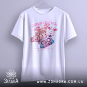 Футболка давай гратись, білого кольору, зображена з фронтального ракурсу, підвішена на вішалці, фон світло-сірий, принт з чотирма мультяшними тваринами та касетою, текст 'давай гратись' виконаний рожевим кольором.