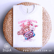 Футболка давай гратись, білого кольору, зображена зверху, складена на плетеній кошику, фон білий, принт з чотирма мультяшними тваринами та касетою, текст 'давай гратись' виконаний рожевим кольором.