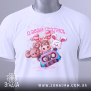 Футболка давай гратись, білого кольору, зображена зверху, складена на поверхні, фон світло-сірий, принт з чотирма мультяшними тваринами та касетою, текст 'давай гратись' виконаний рожевим кольором, на футболці є етикетка з зеленим шнурком.