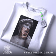 Футболка Давид арт, біла, складена на поверхні, фон світло-сірий, принт зображає статую Давида з язиком, що висунений, та рукою, що торкається обличчя, з етикеткою на шнурку.