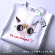 Футболка де моя кавуся. Біла футболка з принтом кота в окулярах, зображена зверху на столі. Принт чіткий, текст червоним кольором, на футболці є етикетка з зеленим шнурком.