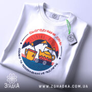 Футболка день обміну не підлягає. Біла футболка з округлим вирізом, зображена з верхнього ракурсу. На фоні світло-сірий колір. Принт з кольоровими елементами, включаючи чайник і чашку, розташовані в центрі.