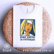 Футболка дякую за п`ятницю, біла, складена на плетеній кошику, фон білий, принт зображає жінку в хустці, яка тримає палець на губах, кольори принта жовтий, синій та оранжевий.