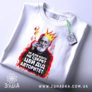 Футболка дід авторитет, біла, зображена з верхнього ракурсу, на фоні світло-сірого градієнта, принт з зображенням чоловіка з бородою, короною та червоними вогнями, текст на українській мові, з зеленим ярликом.