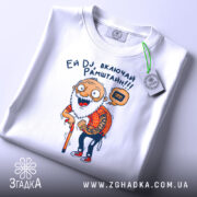 Футболка дід хоче Рамштайн, біла, складена на столі, фон світло-сірий, принт з дідусем у червоній сорочці в клітинку, який тримає палицю, з етикеткою на комірці, текст 'Ей DJ, включай Рамштайн!!!'.