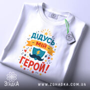 Футболка дідусь мій герой, біла, складена на столі, фон світло-сірий, принт кольоровий з написом 'ДІДУСЬ МІЙ ГЕРОЙ!' та зображенням щита і блискавки, з етикеткою.