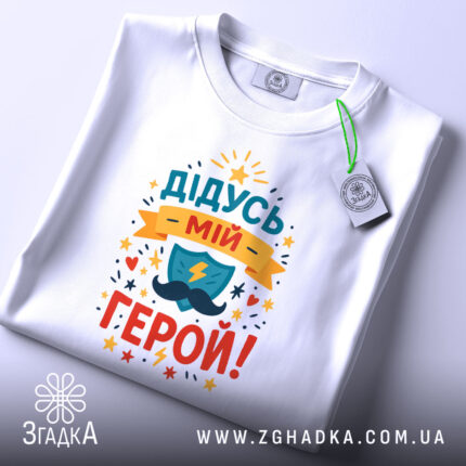 Футболка дідусь мій герой, біла, складена на столі, фон світло-сірий, принт кольоровий з написом 'ДІДУСЬ МІЙ ГЕРОЙ!' та зображенням щита і блискавки, з етикеткою.