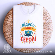 Футболка дідусь мій герой, біла, складена на плетеній кошику, фон світло-сірий, принт кольоровий з написом 'ДІДУСЬ МІЙ ГЕРОЙ!' та зображенням щита і блискавки.