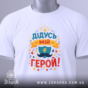 Футболка дідусь мій герой, біла, складена на столі, фон світло-сірий, принт кольоровий з написом 'ДІДУСЬ МІЙ ГЕРОЙ!' та зображенням щита і блискавки, з етикеткою.