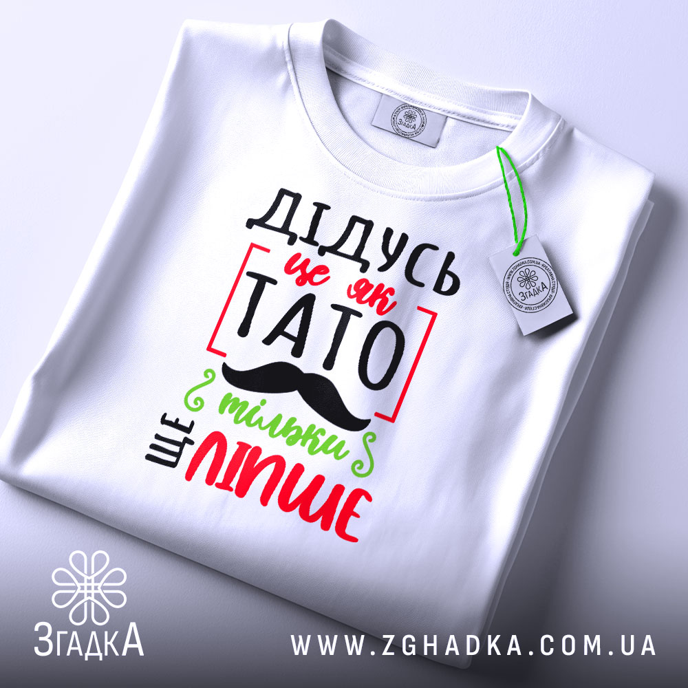 Футболка дідусь це як тато, біла, зображена зверху, на столі, фон світло-сірий, принт з червоним, зеленим і чорним текстом, що говорить 'ДІДУСЬ ЦЕ ЯК ТАТО ТІЛЬКИ ЩЕ ЛІПШЕ', з етикеткою на зеленій стрічці.