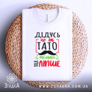 Футболка дідусь це як тато, біла, зображена зверху, на плетеній кошику, фон світлий, принт з червоним, зеленим і чорним текстом, що говорить 'ДІДУСЬ ЦЕ ЯК ТАТО ТІЛЬКИ ЩЕ ЛІПШЕ'.
