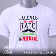 Футболка дідусь це як тато, біла, зображена зверху, на столі, фон світло-сірий, принт з червоним, зеленим і чорним текстом, що говорить 'ДІДУСЬ ЦЕ ЯК ТАТО ТІЛЬКИ ЩЕ ЛІПШЕ', з етикеткою на зеленій стрічці.