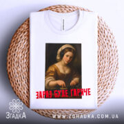 Футболка діджей дівчина, біла, складена на плетеній основі, фон світлий, принт з портретом дівчини в навушниках, текст червоним шрифтом 'ЗАРАЗ БУДЕ ГАРЯЧЕ'.