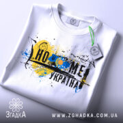 Футболка дім Україна, біла, зображена на столі, фон світлий, принт з жовто-блакитними кольорами, прикріплена етикетка з зеленим шнурком.