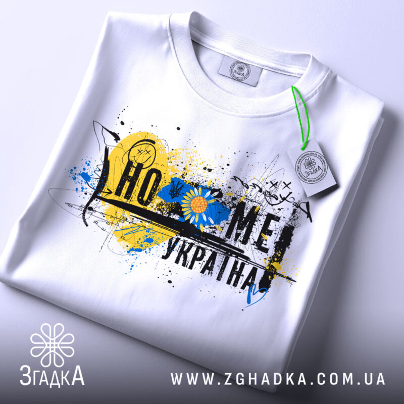 Футболка дім Україна, біла, зображена на столі, фон світлий, принт з жовто-блакитними кольорами, прикріплена етикетка з зеленим шнурком.