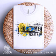 Футболка дім Україна, біла, складена на плетеній основі, фон нейтральний, принт містить жовто-блакитний колір, зображення соняшника та графічні елементи.