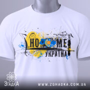 Футболка дім Україна, біла, зображена на столі, фон світлий, принт з жовто-блакитними кольорами, прикріплена етикетка з зеленим шнурком.