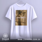 Футболка дівчата можуть бути білого кольору, підвішена на вішалці з нейтральним фоном. Принт з зображенням жінок та текстом "Girls Will Be Girls" чітко видимий на передній частині футболки.