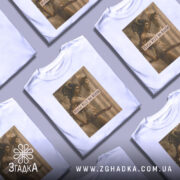 Футболка дівчата можуть бути білого кольору, з прямим кроєм, на манекені з правого боку. На футболці розташований принт з зображенням двох жінок, одна з яких тримає меч, а текст "Girls Will Be Girls" розміщений на фоні.