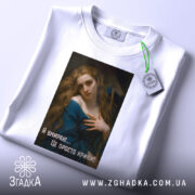 Футболка дівчина кринж, біла футболка, складена на поверхні, принт з дівчиною у блакитному платті, текст 'Я вмираю... Це просто кринж!' на чорному фоні, зелена етикетка на комірці, фон світлий.