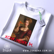 Футболка дівчина піца, біла, зображена на столі, фон світло-сірий, принт з портретом жінки в червоній сукні, яка тримає шматок піци, текст "Хочу, можу, їм" червоним шрифтом, з етикеткою на комірці.