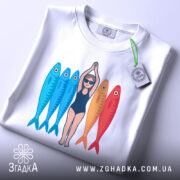 Футболка дівчина рибки білого кольору, зображена зверху, на ній видно принт з дівчиною в купальнику та рибками, які мають яскраві кольори: блакитний та червоний, на футболці є етикетка з зеленим шнурком.