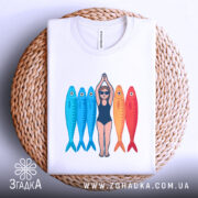 Футболка дівчина рибки білого кольору, зображена зверху на плетеній кошику, фон світлий, принт з дівчиною в купальнику та рибками, які мають блакитний та червоний кольори, футболка акуратно складена.