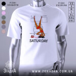 Футболка дівчина субота, біла, зображена з фронтального ракурсу, на фоні світло-сірого кольору, принт з дівчиною у червоному купальнику, що висить вниз головою, з телефоном на столі та написом 'SATURDAY' під малюнком.