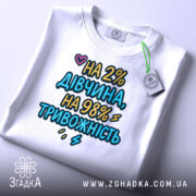 Футболка дівчина тривожність подарунок. Біла футболка, складена на столі, з текстом 'НА 2% ДІВЧИНА, НА 98% ТРИВОЖНІСТЬ'. Принт яскравий, з сердечком. На футболці є етикетка.