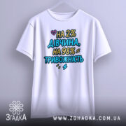 Футболка дівчина тривожність подарунок. Біла футболка, підвішена на вішалці, з кольоровим принтом на грудях. Фон світло-сірий, акцент на тексті.