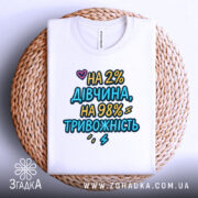 Футболка дівчина тривожність подарунок. Біла футболка, складена на круглому кошику, з принтом 'НА 2% ДІВЧИНА, НА 98% ТРИВОЖНІСТЬ'. Фон світлий, акцент на тексті.