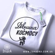 Футболка дівчина з космосу, біла, складена на поверхні, фон світло-сірий, принт чорний з написом 'Дівчина з космосу' та малюнком астронавта, на комірці є етикетка.