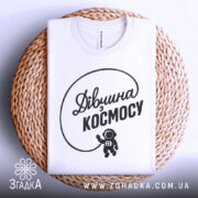 Футболка дівчина з космосу, біла, складена на плетеній кошику, фон білий, принт чорний з написом 'Дівчина з космосу' та малюнком астронавта.
