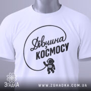 Футболка дівчина з космосу, біла, складена на поверхні, фон світло-сірий, принт чорний з написом 'Дівчина з космосу' та малюнком астронавта, на комірці є етикетка.