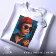 Футболка дівчина з пістолетом, біла, з верхнього ракурсу, на фоні світло-сірого кольору, принт з зображенням дівчини в червоній шапці та чорних окулярах, пістолет у руці, з етикеткою на шнурку.