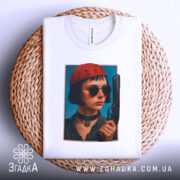 Футболка дівчина з пістолетом, біла, з верхнього ракурсу, на фоні плетеного кошика, принт з зображенням дівчини в червоній шапці та чорних окулярах, пістолет у руці.