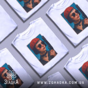 Футболка дівчина з пістолетом, біла, з фронтального ракурсу, на фоні світло-сірого кольору, принт з зображенням дівчини в червоній шапці та чорних окулярах, тримає пістолет.