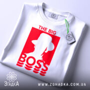 Футболка для босса, білого кольору, складена на столі, фон світло-сірий, принт червоного кольору з написом 'THE BIG BOSS' та силуетом людини в капелюсі, на футболці прикріплена етикетка.