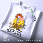 Футболка для електрика білого кольору, зображена зверху, на ній видно етикетку з зеленим шнурком, фон світло-сірий, принт з мультяшним персонажем з червоним волоссям, що тримає електричний вимикач, текст "Trust Me, I'm The ELECTRICIAN" під малюнком.