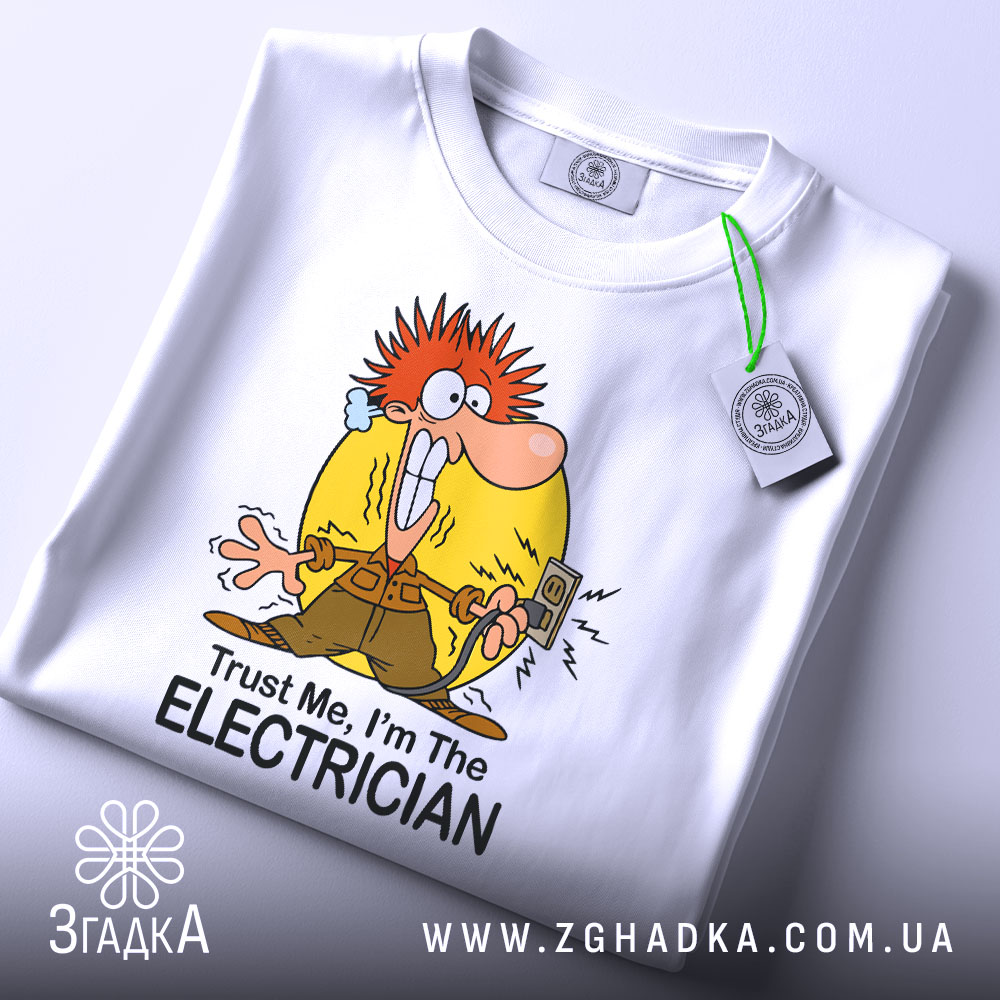 Футболка для електрика білого кольору, зображена зверху, на ній видно етикетку з зеленим шнурком, фон світло-сірий, принт з мультяшним персонажем з червоним волоссям, що тримає електричний вимикач, текст "Trust Me, I'm The ELECTRICIAN" під малюнком.
