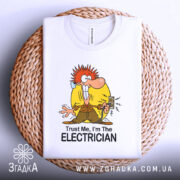 Футболка для електрика білого кольору, зображена зверху на плетеному кошику, фон білий, принт з мультяшним персонажем з червоним волоссям, що тримає електричний вимикач, текст "Trust Me, I'm The ELECTRICIAN" під малюнком.