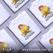 Футболка для електрика білого кольору, зображена з правого ракурсу на манекені, фон світло-сірий, принт з мультяшним персонажем з червоним волоссям, що тримає електричний вимикач, текст "Trust Me, I'm The ELECTRICIAN" під малюнком.