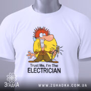 Футболка для електрика білого кольору, зображена зверху, на ній видно етикетку з зеленим шнурком, фон світло-сірий, принт з мультяшним персонажем з червоним волоссям, що тримає електричний вимикач, текст "Trust Me, I'm The ELECTRICIAN" під малюнком.