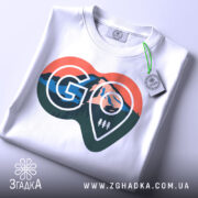 Футболка для мандрівників. Біла футболка, складена на поверхні, з видимим принтом у формі серця. Принт містить зображення гір, лісу та логотип 'GO'. На футболці висять етикетки, фон світлий і чистий.