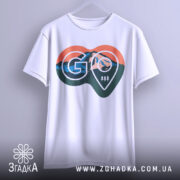 Футболка для мандрівників. Біла футболка, підвішена на вішалці, з фронтального ракурсу. Принт у формі серця з горами та логотипом 'GO' займає центральну частину. Фон світлий, що робить акцент на дизайні футболки.