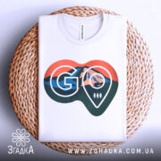 Футболка для мандрівників. Біла футболка, складена на плетеній кошику, з видимим принтом у формі серця. Принт містить зображення гір, лісу та логотип 'GO'. Фон світлий, а кошик додає текстурності до зображення.