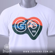 Футболка для мандрівників. Біла футболка, складена на поверхні, з видимим принтом у формі серця. Принт містить зображення гір, лісу та логотип 'GO'. На футболці висять етикетки, фон світлий і чистий.