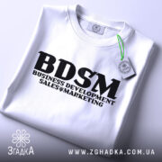 Футболка для маркетолога, білого кольору, складена на столі, з видимим етикеткою зеленого кольору, принт чорного кольору з написом 'BDSM BUSINESS DEVELOPMENT SALES & MARKETING'.