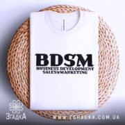 Футболка для маркетолога, білого кольору, складена на плетеній кошику, фон білий, принт чорного кольору з написом 'BDSM BUSINESS DEVELOPMENT SALES & MARKETING'.