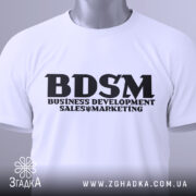Футболка для маркетолога, білого кольору, складена на столі, з видимим етикеткою зеленого кольору, принт чорного кольору з написом 'BDSM BUSINESS DEVELOPMENT SALES & MARKETING'.
